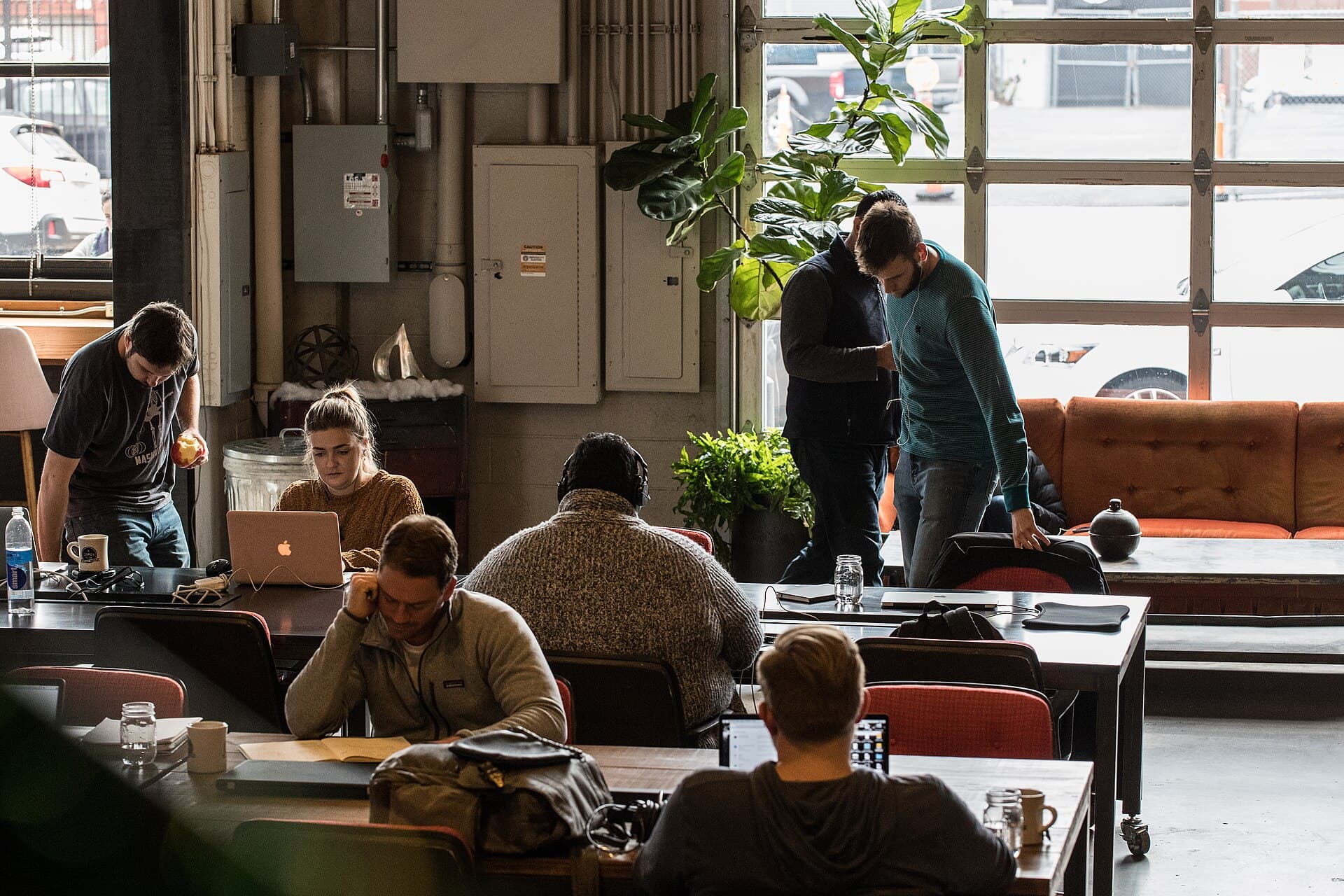 A busy coworking space with people working at laptops beside a big window